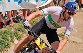 Imagem: Fotografia. Um homem, vestindo camiseta branca com a bandeira do Brasil, capacete azul, óculos de sol e luvas, está em uma bicicleta preta com uma placa de identificação com o número 3. Ao fundo, barro, grama e parte do corpo de pessoas.   Fim da imagem.