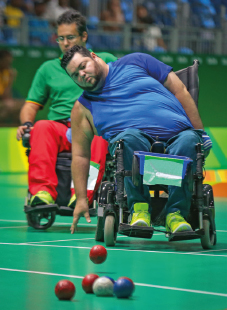 Imagem: Fotografia. Um homem, de camisa azul e calça verde, sentado em uma cadeira de rodas, com o braço direito esticado na direção do chão e a mão aberta, olha para uma bola vermelha. À frente dele, outras bolas estão posicionadas no chão. Ao fundo, outro homem com camisa verde e calça vermelha, e uma arquibancada.  Fim da imagem.