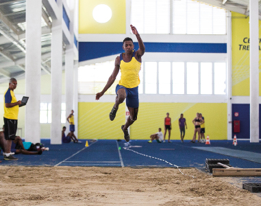 Imagem: Fotografia. Ao centro, um homem com regata amarela e bermuda azul salta na areia com a perna direita dobrada e a esquerda esticada. Os braços estão esticados. Atrás, outras pessoas observam. Ao fundo, pilares brancos e paredes amarelas.  Fim da imagem.