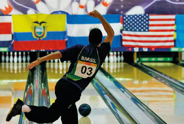 Imagem: Fotografia. Um homem, com uma camiseta em tons de verde escrito BRASIL e com uma placa de identificação com o número 3, lança uma bola de boliche. A perna direita está levantada e o braço direito estendido para cima. Ao fundo, uma pista de madeira com pinos de boliche brancos. Há bandeiras do Equador, Guatemala e Estados Unidos.   Fim da imagem.