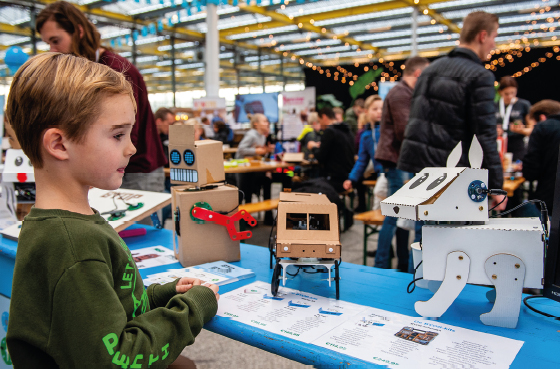 Imagem: Fotografia. À esquerda, um menino vestindo blusa verde, olha para uma mesa azul. Sobre a mesa há um robô marrom com braço vermelho com formato de chave de fenda, um carro marrom com rodas pretas, e um cachorro branco com formato geométrico. Ao fundo, mesas e pessoas. Fim da imagem.