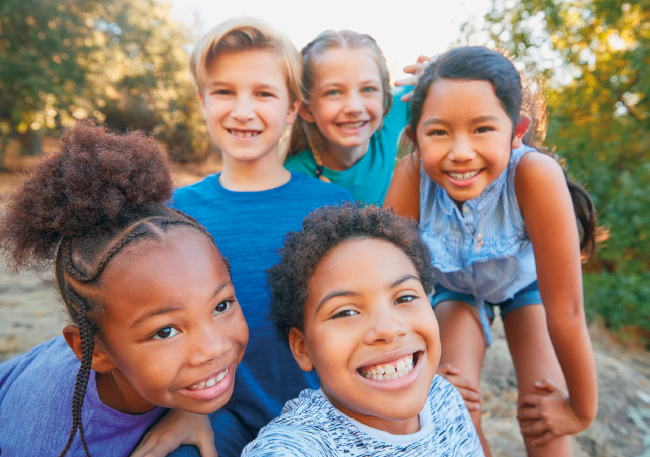 Imagem: Fotografia. Destaque para cinco crianças que sorriem. Ao fundo, árvores. Fim da imagem.