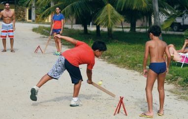 Imagem: Fotografia. Na frente, dois meninos, um com camiseta laranja e bermuda azul, segura um taco e aponta na direção de uma bola amarela, o outro menino olha. No fundo, uma mulher segura um taco e, ao lado dela, há um homem e ambos olham a bola. Fim da imagem.