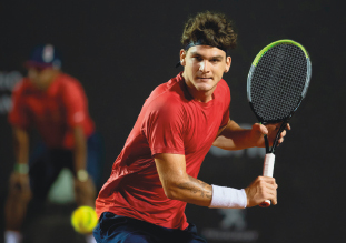 Imagem: Fotografia. Um homem vestindo camiseta vermelha e com faixa branca nos pulsos, segura uma raquete de tênis e olha para uma bola verde.  Fim da imagem.