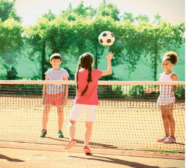 Imagem: Fotografia. Uma criança, usando cabelo trançado, vestindo camiseta vermelha e bermuda branca, está com os braços dobradas, mãos abertas e olha uma bola que está no alto. À frente dela, há uma rede com borda branca rente ao chão. Do outro lado da rede, duas crianças olham para a bola.  Fim da imagem.