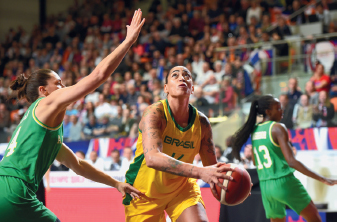 Imagem: Fotografia. No centro, há uma mulher vestindo uma regata amarela escrito BRASIL. Ela está segurando uma bola laranja nas mãos e olha para cima. À direita dela, há outra mulher, vestindo regata verde, que está com o braço direito erguido. Ao fundo, pessoas sentadas na arquibancada.  Fim da imagem.