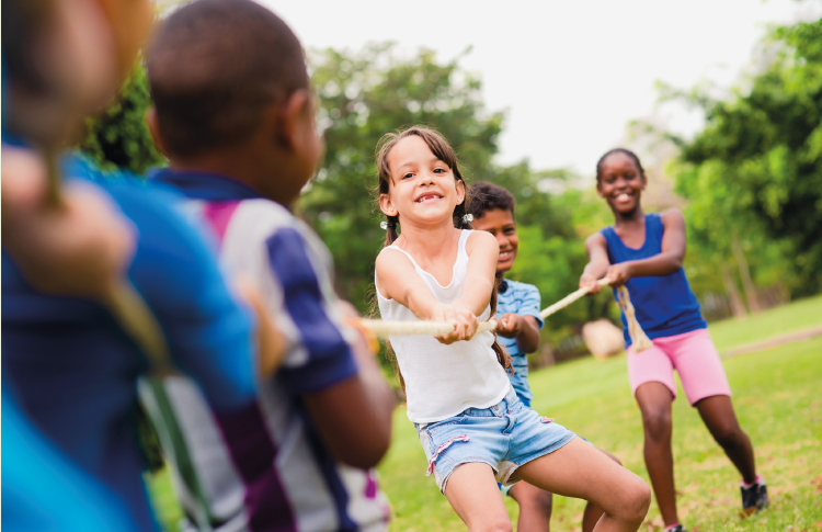 Imagem: Fotografia. No centro, destaque para uma garota com cabelo preso em maria-chiquinha, vestindo regata branca e bermuda jeans, está com pernas dobradas, braços estendidos e segura uma corda. Atrás e na frente dela, mais crianças seguram a corda.  Fim da imagem.