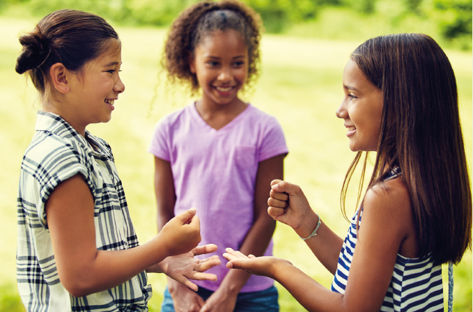 Imagem: Fotografia. Na frente, destaque para duas crianças com os braços dobrados, uma mão aberta e uma mão fechada, à frente do corpo. Atrás, uma menina está sorrindo.  Fim da imagem.