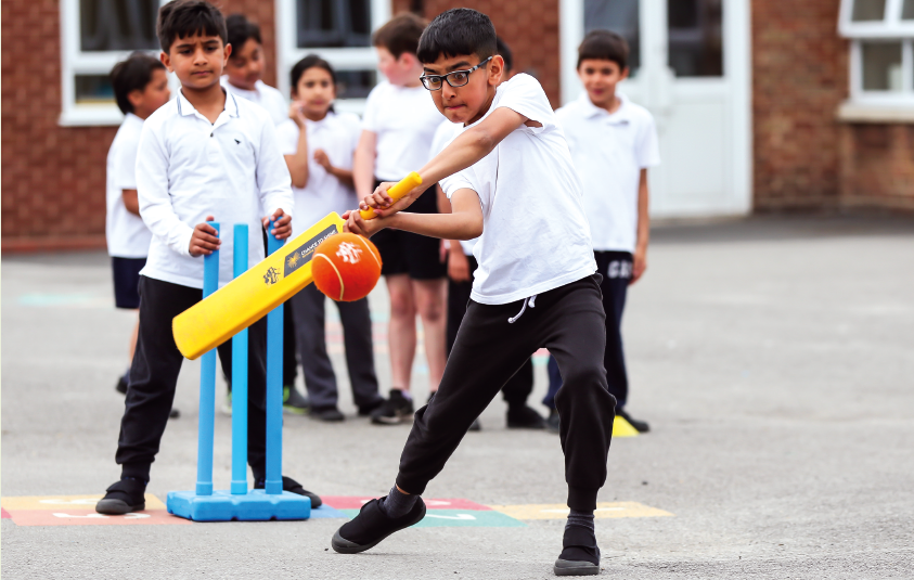Imagem: Fotografia. Um menino vestindo uma camiseta branca e calça preta segura um taco amarelo com as duas mãos e aponta para uma bola laranja. Seus lábios estão contraídos e o cenho franzido. Ao fundo, um objeto com base retangular e três hastes na vertical. Ao redor, crianças em fila.  Fim da imagem.
