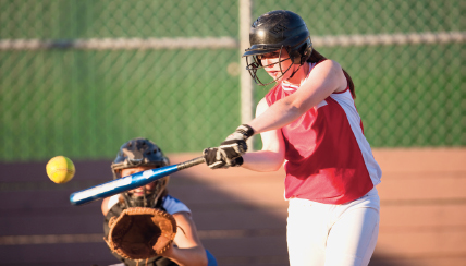 Imagem: Fotografia. Na frente, uma criança de capacete segura um taco com as duas mãos e aponta para uma bola amarela. Ao fundo, uma criança de capacete e luva está abaixada e com a mão na frente do corpo.  Fim da imagem.