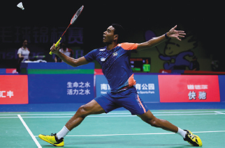 Imagem: Fotografia. Destaque para um homem, com cabelo curto e vestindo camiseta azul. Ele está com as pernas esticadas e, com o braço direito dobrado, segura uma raquete e aponta para uma bola. Fim da imagem.