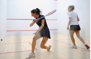 Imagem: Fotografia. Duas mulheres, vestindo camiseta e saia, seguram uma raquete e olham para uma bola preta. Elas estão dentro de uma sala branca com linhas vermelhas e um piso de madeira.  Fim da imagem.