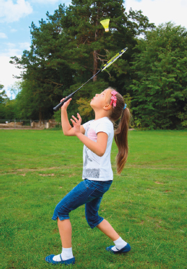 Imagem: Fotografia. Uma menina, com o cabelo preso, vestindo uma camiseta branca e calça jeans, está com os braços dobrados, segura uma raquete e aponta para uma peteca verde. Ao fundo, árvores. Fim da imagem.
