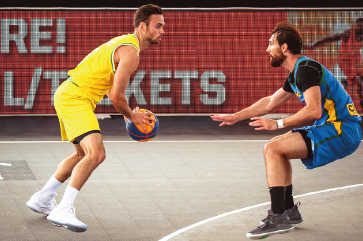 Imagem: Fotografia. À esquerda, um homem, com cabelo curto, vestindo regata e bermuda amarela, está com os braços flexionados e, com as mãos abertas, segura uma bola de basquete. À direita, outro homem, com regata e bermuda azul, está com os joelhos dobrados e os braços esticados para frente.  Fim da imagem.