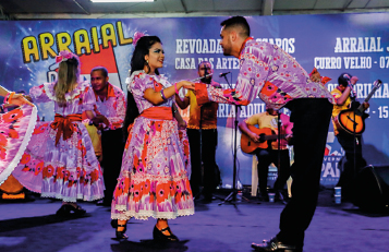 Imagem: Fotografia. No centro, um homem, vestindo uma camiseta florida roxa e calça preta, está com o tronco inclinado para frente e o braço dobrado na direção de uma mulher. A mulher usa um vestido roxo florido e está com o braço dobrado na direção da mão do homem.  Fim da imagem.