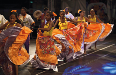 Imagem: Fotografia. Mulheres, em fileira, usam um vestido em tons de amarelo e laranja, e com as mãos seguram a ponta da saia.  Ao fundo, homens com chapéu e camiseta cinza.  Fim da imagem.