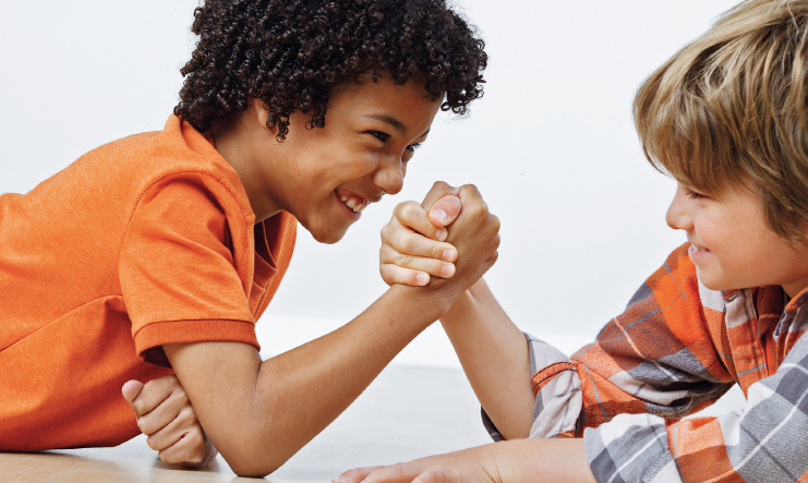 Imagem: Fotografia. Destaque para dois meninos, de frente um para o outro, apoiando o cotovelo em uma superfície e encaixando as mãos de lado. Eles estão com o cenho franzido e sorriem.  Fim da imagem.