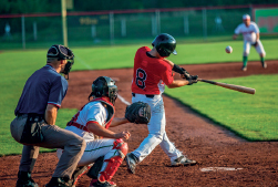 Imagem: Fotografia. No centro, uma pessoa vestindo camiseta vermelha e capacete preto, segura com as duas mãos um taco e aponta para uma bola branca. Atrás dele, duas pessoas com capacete e luvas olham a bola.  Fim da imagem.