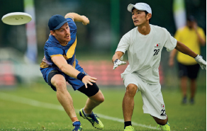 Imagem: Fotografia. À direita, um homem vestindo camiseta, luvas e chapéu branco, está agachado no chão, com os braços inclinados, as mãos abertas e observa um disco branco. À esquerda, um homem com camisa e chapéu azul está com o braço esticado para frente.  Fim da imagem.