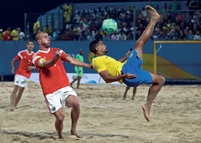 Imagem: Fotografia. No centro, um homem vestindo camiseta amarela e bermuda azul, está com a perna direita para cima e olha para uma bola verde. Ao lado dele, um homem usando camiseta vermelha, está com os olhos fechados, boca contraída e com os braços esticados para frente. O chão é de areia e ao fundo há pessoas em uma arquibancada.  Fim da imagem.
