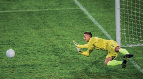 Imagem: Fotografia. Um homem está deitado na grama e com os braços flexionados para frente, na direção de uma bola. O chão é de grama e ao fundo há uma trave branca.  Fim da imagem.