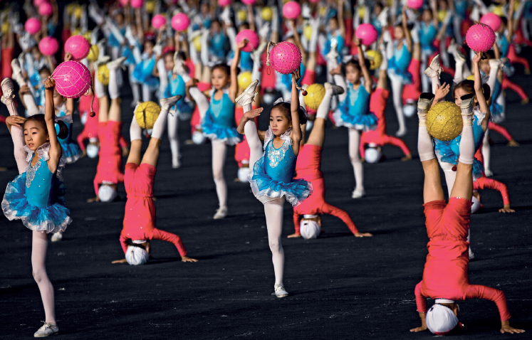 Imagem: Fotografia. Diversas meninas usando um vestido azul, meia alta branca, estão segurando uma bola rosa com a mão esquerda e com a mão direita seguram a perna levantada na altura da cabeça. Entre elas, há diversas crianças com roupa vermelha e touca branca, de ponta cabeça, com as mãos apoiadas no chão e com uma bola amarela entre as pernas que estão para cima. Fim da imagem.