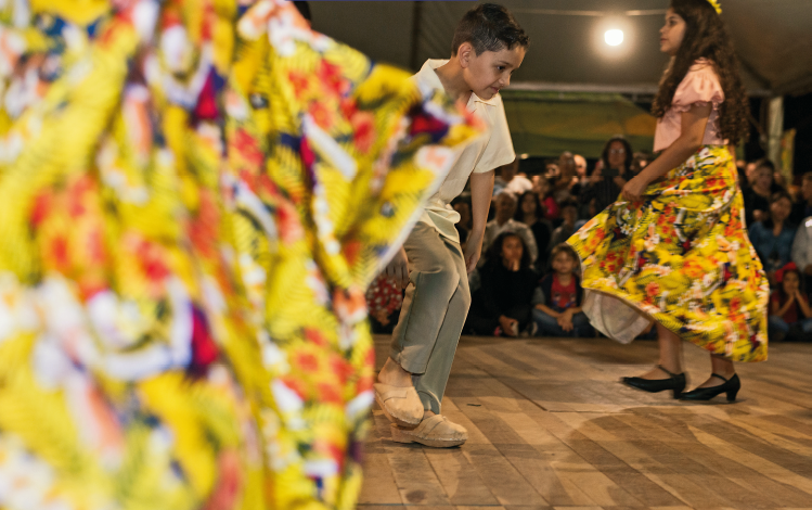 Imagem: Fotografia. No centro, um menino com cabelo curto, vestindo camiseta branca e calça cinza, está com o tronco inclinado para frente. Na sua frente uma menina com cabelo longo, vestindo camiseta rosa e saia amarela florida, segura a ponta da saia. Ao fundo, pessoas sentadas observam.  Fim da imagem.