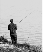 Imagem: Fotografia em preto e branco. Um homem de costas nas margens de um rio, segurando uma vara de pescar.  Fim da imagem.