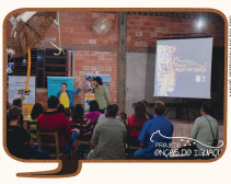 Imagem: fotografia de pessoas sentadas em cadeiras de costas olhando para uma projeção. No fundo duas pessoas em pé apontando para o telão.  Fim da imagem.