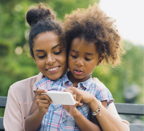 Imagem: Fotografia. Uma mulher com cabelo preso e camiseta rosa está sorrindo e com os braços em volta de uma menina com cabelo encaracolado e camiseta xadrez. A menina está mexendo em um aparelho eletrônico e a mulher está ajudando. Ao fundo, árvores. Fim da imagem.