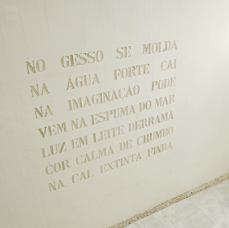 Imagem: Fotografia. Uma parede branca com o texto:  NO GESSO SE MOLDA  NA ÁGUA FORTE CAI  NA IMAGINAÇÃO PODE  VEM NA ESPUMA DO MAR  LUZ EM LEITE DERRAMA  COR CALMA DE CHUMBO  DE CAL EXTINTA FINDA.  Fim da imagem.