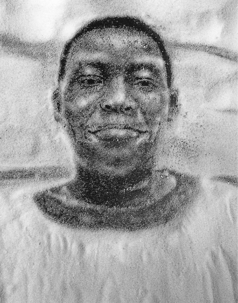 Imagem: Pintura em preto e branco composta por grãos em tons de preto. Rosto de um jovem com cabelo curto, olhos e nariz grandes e lábios grossos. Fim da imagem.