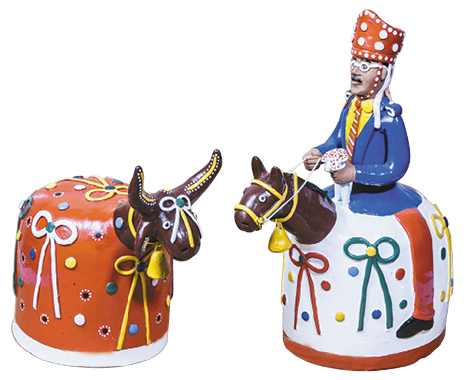 Imagem: Fotografia. À direita, boneco de um homem com chapéu laranja, óculos e terno azul. Em volta dele há um cavalo-marinho colorido. À esquerda, boneco de um bumba meu boi colorido. Fim da imagem.