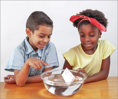 Imagem: Fotografia. Duas crianças observando um papel em formato de barco sobre uma vasilha de água. Menino de cabelo curto castanho, vestindo camiseta azul. Ao lado, menina de cabelo cacheado castanho e faixa vermelha, vestindo camiseta amarela. Fim da imagem.