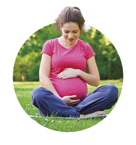 Imagem: Fotografia. Mulher grávida de cabelo longo castanho, vestindo camiseta rosa e calça azul. Ela está com as mãos apoiadas na barriga, como se estivesse segurando-a. Fim da imagem.