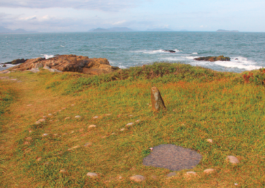 Imagem: Fotografia. Vista de campo com gnômon formada por pedra maior e semicírculo formando por pedras menores. No horizonte da imagem está o mar. Fim da imagem.