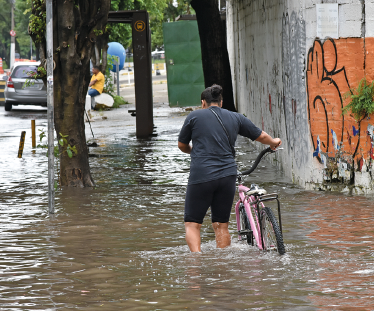Imagem: Fotografia. Destaque de uma mulher de cabelo curto preto, vestindo camiseta e bermuda preta. Está levando uma bicicleta por uma calçada alagada com água até a altura do joelho. Fim da imagem.