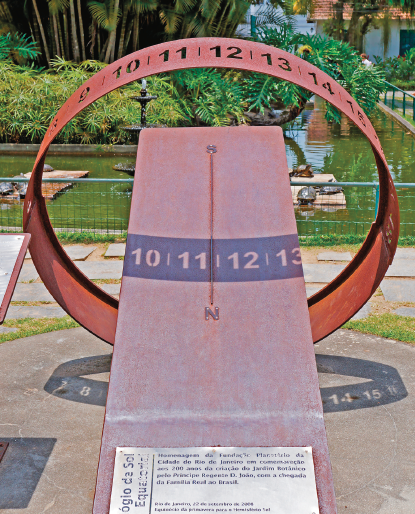 Imagem: Fotografia. Relógio do sol com linha no centro de uma barra retangular. Ao redor, um círculo de ferra oco transpassando pela barra. Fim da imagem.