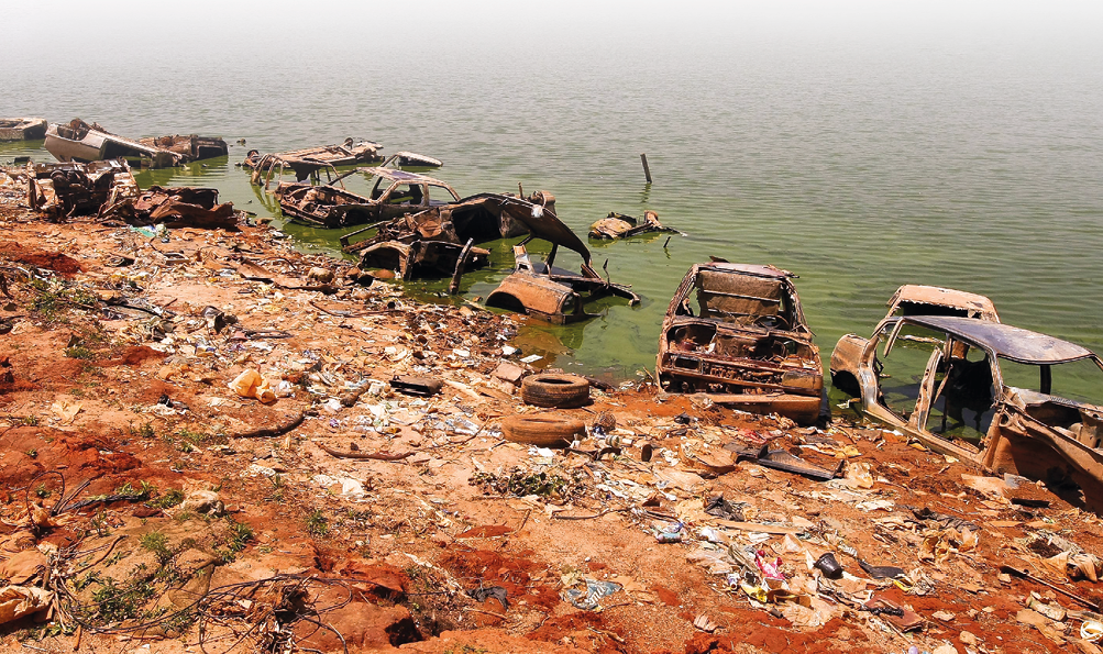 Imagem: Fotografia. Vista de margem do mar com lixos e dejetos. Sobre a água há carcaças de carros enferrujados. Fim da imagem.