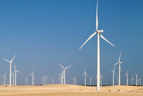 Imagem: Fotografia. Campo desértico com muitos aerogeradores em formato de cata-vento. Fim da imagem.