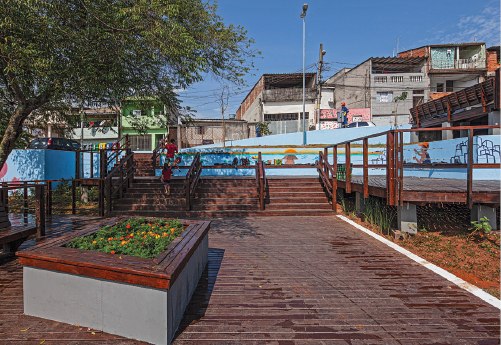 Imagem: Fotografia. Vita de praça coberta por chão de madeira com flores sobre vasos e árvores. Atrás da praça há casas.   Fim da imagem.