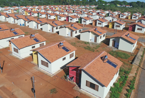 Imagem: Fotografia. Vista aérea de casas baixas iguais em longas fileiras lado a lado em cidade. Sobre o telhado há placas de coleta de luz solar.  Fim da imagem.