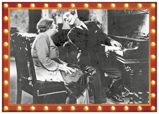 Imagem: 1927 - O cantor de jazz é o primeiro filme com trechos com diálogos sonoros.  Foto em preto e branco. Um homem com terno e gravata está sentado com as mãos sobre um piano e com a cabeça inclinada para o lado. Em seguida, uma mulher com vestido está sentada e com a cabeça inclinada para o lado.  LEGENDA: Cena do filme O cantor de jazz, dirigido por Alan Crosland, de 1927. FIM DA LEGENDA.   Fim da imagem.