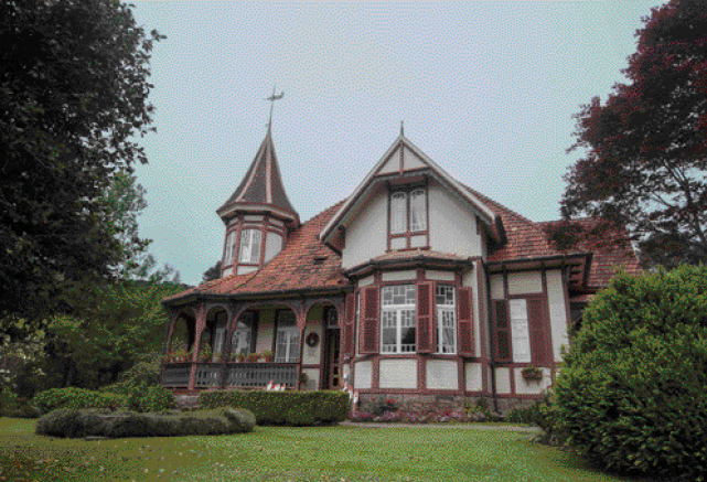 Imagem: Fotografia. Casa grande com paredes bege e detalhes marrons. Na lateral há uma torre com telhado pontudo e em volta, plantas e árvores.  Fim da imagem.