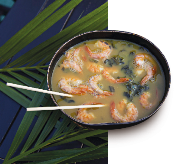 Imagem: Fotografia. Uma tigela preta e dentro há uma sopa com algas e camarão. Ao fundo, folhas grandes e finas.  Fim da imagem.