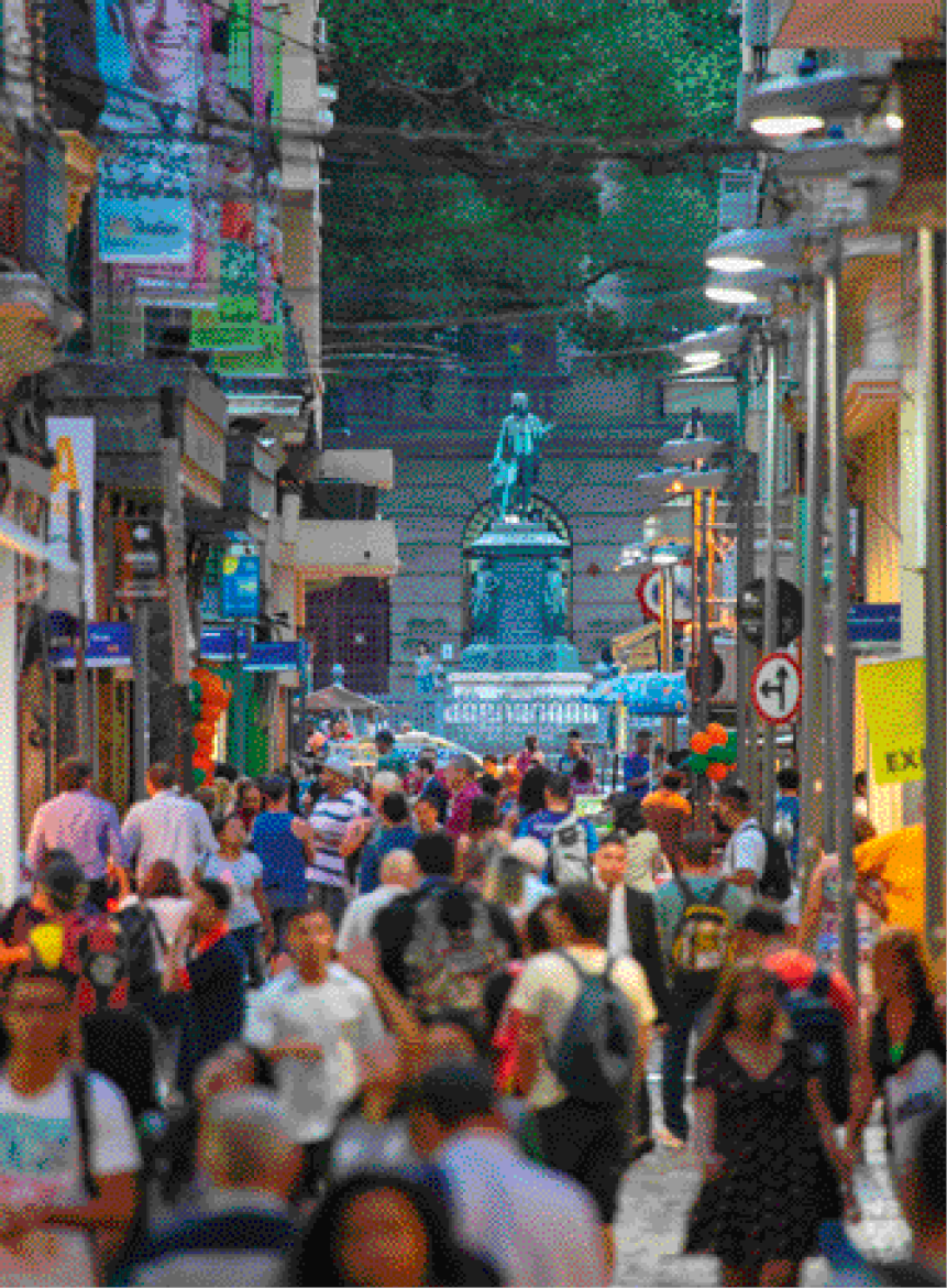 Imagem: Fotografia. Várias pessoas andando em uma rua. Nas laterais há lojas, postes com as luzes acesas, placas e faixas. Ao fundo, um monumento na frente de uma construção grande. Fim da imagem.