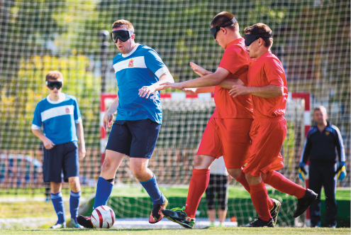 Imagem: Fotografia. Dois homens com uniformes azuis e máscara de olhos. Um deles está com uma bola próximo aos pés. À direita, dois homens de uniforme vermelho indo em direção ao homem que está com a bola.  Fim da imagem.