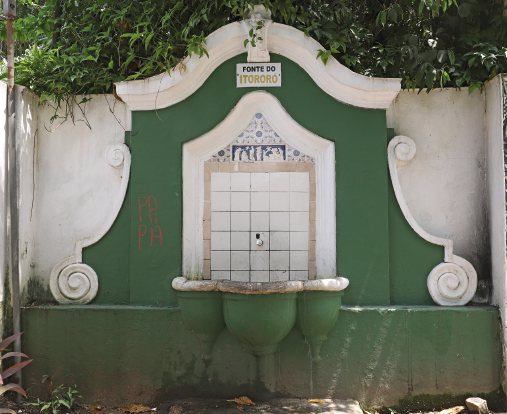 Imagem: Fotografia. Uma fonte com uma pia verde. No fundo, vegetação. Fim da imagem.