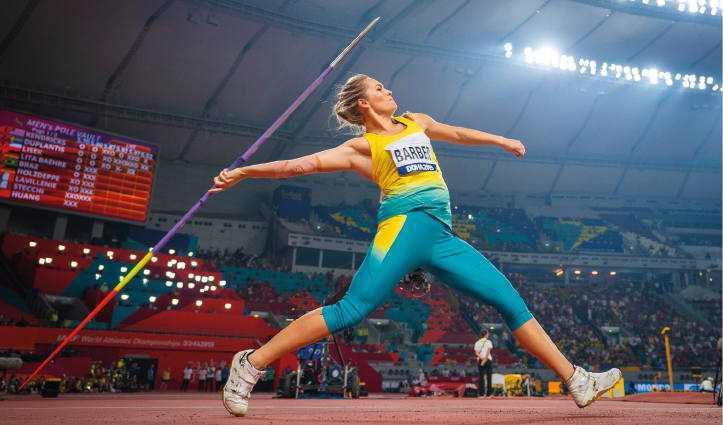 Imagem: Fotografia. Uma mulher com cabelos loiros e presos, usando uniforme verde e amarelo. Ela está de perfil segurando um dardo (objeto em forma de lança). Fim da imagem.