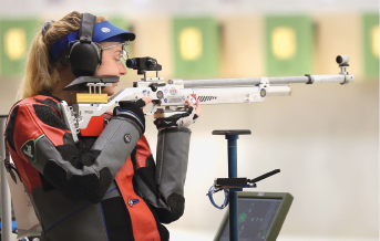 Imagem: Fotografia. Uma mulher com cabelos lisos, usando fone de ouvido, viseira e segurando com as mãos uma espingarda. Fim da imagem.